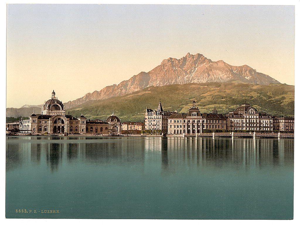 The image depicts a picturesque lakeside scene with classical European architecture in the foreground. A prominent structure on the left features an ornate facade and dome, suggesting it could be of historical significance or serve as a public building like a station or town hall. To its right are several multi-storied buildings with traditional design elements such as steeply pitched roofs, large windows, and decorative cornices.

In the middle ground, we see calm blue-green waters reflecting the architectural ensemble and hints of greenery along the lake's edge. The background is dominated by a majestic mountain range under a clear sky, which adds to the serene atmosphere of the scene.

The warm colors suggest that this image was taken during golden hour or has been edited for an antique aesthetic effect. A handwritten inscription at the bottom left corner indicates "Lucerne," identifying one possible location in Switzerland where such scenes might be found. This photograph could serve as a postcard, capturing the beauty and charm of Swiss architecture and landscape.

The overall composition conveys tranquility and historical elegance, potentially appealing to those interested in European travel or history.