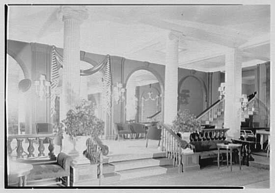 The image depicts an elegant and spacious ballroom from the early 20th century. The room is adorned with a series of tall, white columns that create an imposing archway leading to another part of the building. American flags are prominently displayed on both sides of this entrance.

In the foreground, there's a long staircase with ornate balustrades and wooden steps. At the foot of the stairs, we see two large chairs upholstered in dark fabric, which contrast against the lighter tones of the room.

To the left side, near one of these columns, stands an exquisite urn-style vase containing lush green foliage that adds to the opulence of this setting. On a raised platform towards the backside lies another elaborate vase filled with flowers, enhancing the grandeur of the space.

The walls are decorated with framed artworks and ornate wall sconces provide lighting along one side. A large window or possibly glass doors on the right allow natural light to filter through, illuminating this luxurious interior.

Overall, the ballroom exudes a sense of historical elegance and is reminiscent of social gatherings in affluent households during that era.