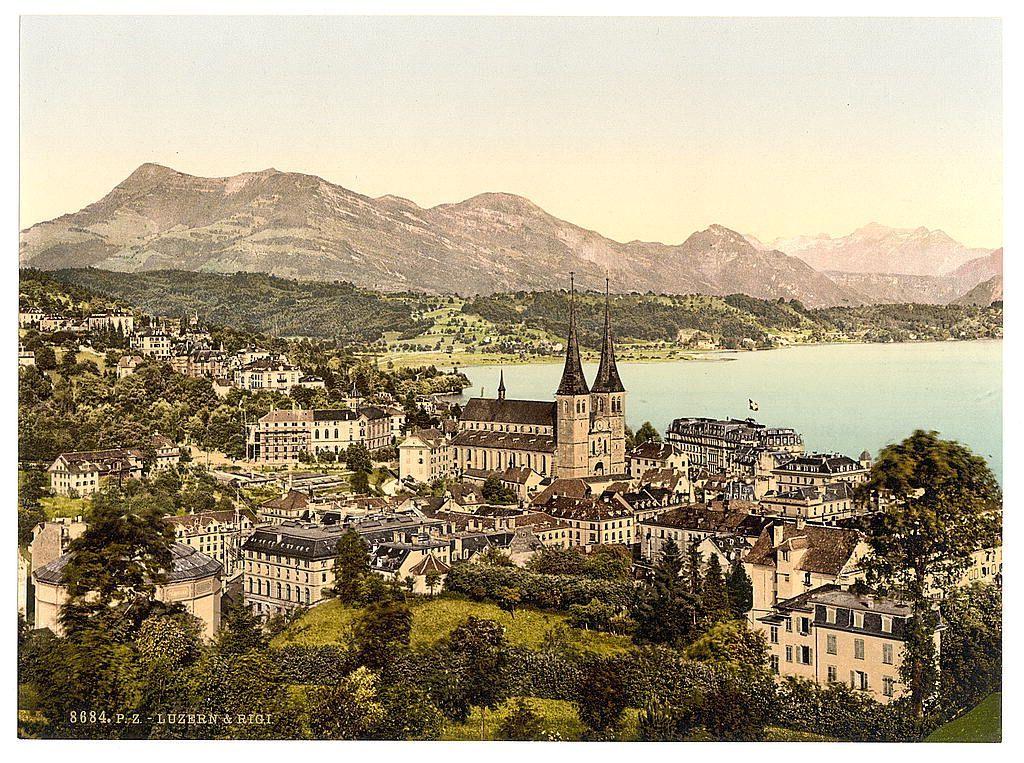 The image depicts a picturesque town set against the backdrop of mountains and water. Dominating the center is an ornate church with two prominent spires, indicative of Gothic architecture, its facade adorned with detailed stonework and surrounded by smaller buildings in various architectural styles suggesting historical significance. The surrounding structures are densely packed within what appears to be narrow streets, typical of European town planning from a past era.

The landscape beyond the town is lush with greenery, hinting at fertile plains or rolling hills that contribute to its scenic value. In the distance, gentle mountains rise above the horizon line, partially shrouded in light mist, which adds depth and serenity to the overall scene. The colors of the image are somewhat muted yet warm, likely due to the photograph's age, with shades ranging from yellowish-brown for the buildings to green for vegetation.

The presence of text at the bottom indicates that this image is a historical print or postcard; however, specific details such as size and condition cannot be discerned without further context. The overall impression conveyed by the visual elements suggests a peaceful yet vibrant town in Switzerland during its formative years.