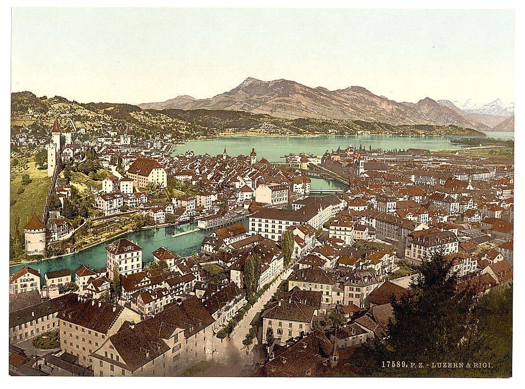 The image displays a panoramic view of an old European town with dense clusters of buildings featuring steep, gabled roofs and red-tiled facades. The architecture suggests this could be somewhere in Switzerland or Italy during the late 19th to early 20th century based on its style.

A prominent river winds through the heart of the settlement, reflecting a golden hue that contrasts with the town's earthy tones. On one side of the waterway stands an imposing castle-like structure with white walls and dark roofing, serving as a focal point in the foreground.

Beyond the urban landscape lies expansive greenery leading up to a range of mountains under a hazy sky. The gentle undulations of these natural formations extend into the distance where they meet another mountainous horizon adorned with snow-dusted peaks, suggesting an alpine climate.

The image is tinted brown and greenish in places, which may be due to age or intentional colorization for artistic effect. A handwritten note at the bottom indicates "17589.P.Z.- LUZERN & RIGI," implying this town could be Luzern with a view towards Rigi—a mountain range known as the 'Swiss Alps'—suggested by the background features.

In terms of context, given that it is associated with Swiss imagery and dated to approximately between 1890-1900, one can infer it was captured during a time when postcards were popular for documenting trave [...]