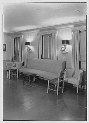 The image is a black and white photograph showing an elegant living room with classic furnishings from 1946. A large, plush sofa takes center stage in the middle of the room, flanked by two armchairs on either side. The furniture appears to be made from light-colored materials such as linen or silk.
The walls are adorned with curtains that hang evenly across tall windows dressed with drapes. These provide a sense of privacy and add warmth to the atmosphere within the space. A couple of wall-mounted lamps, each equipped with two lights emitting warm lighting, enhance this ambiance further by casting soft shadows around the room's features.
On one side of the sofa stands an end table with a lamp on top, presumably serving as a reading or conversation light for guests seated in the living area. The flooring is not fully visible but seems to be a polished wood floor that reflects some of the natural and artificial lighting within the space.
Overall, this setting conveys sophistication and comfort from mid-20th century America's affluent social circles where such elegant interiors would typically belong.