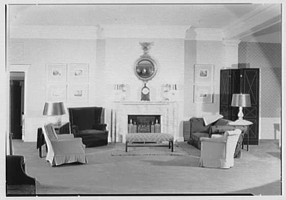 The image depicts a vintage black-and-white photograph of an elegant living room, possibly from the mid-20th century. The space is furnished with classic pieces including armchairs, sofas, and various tables adorned with decorative items such as lamps, vases, and books. Notably, there's a fireplace at one end of the room featuring ornate details and framed by a large mirror above it. On either side of this central feature are tall floor-to-ceiling windows draped with heavy curtains. The walls bear several pieces of art or photographs in frames, contributing to an atmosphere that is both formal and welcoming.

The furniture appears well-worn yet maintained, indicating the room may have been part of a residence rather than a museum display. This particular photo comes from "The Homestead" in Hot Springs, Virginia, dated May 15th, according to its metadata provided by Gottscho-Schleisner Inc., suggesting that it's a historical snapshot capturing an interior space within this landmark hotel or estate.

In terms of visual details such as colors and textures, the image is limited due to its monochromatic nature. However, one can infer from the contrast between light and shadow where objects are placed in relation to natural light coming through the windows that there's a careful attention paid to lighting which enhances certain features within this well-furnished room.

There s [...]