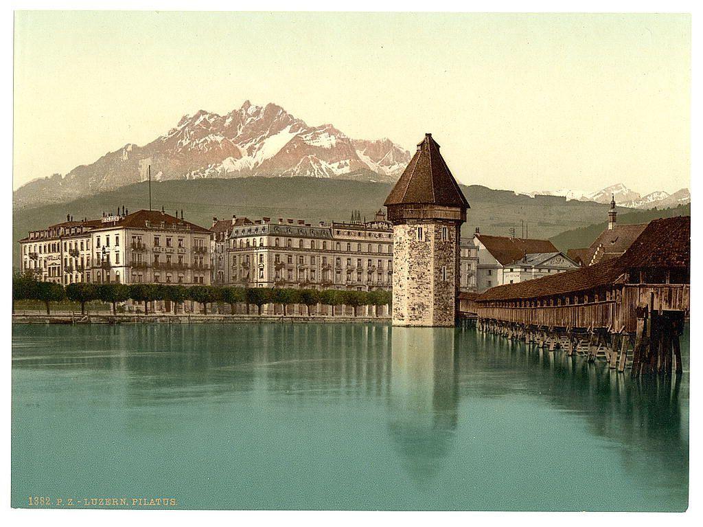 The image presents a picturesque historical scene captured in sepia tones, indicative of an aged photograph. It depicts the iconic Chapel Bridge (Kapellbrücke) spanning across Lake Lucerne with Pilatus mountain majestically looming in the backdrop. The bridge itself is characterized by its Gothic architecture and vibrant red-painted wooden beams that create a striking contrast against the tranquil blue-green waters below.

In addition to the architectural beauty, various buildings flank the sides of the lake, showcasing European design elements such as ornate balconies, dormer windows, and steeply pitched roofs. The waterway reflects these structures, adding depth to the image's composition. This view encapsulates a moment frozen in time, portraying not only the physical attributes but also hinting at the cultural heritage embedded within this Swiss locale.

This specific photograph is marked with "1892 P.Z.-LUZERN PILATUS," suggesting it was taken around that year and capturing Pilatus from Lake Luzern. Such images serve as valuable historical records, allowing us to glimpse into past landscapes and architectural grandeur while appreciating their enduring allure today.