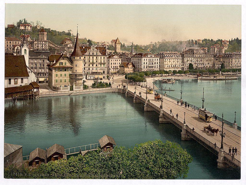 The image depicts a picturesque European riverside scene, likely from the late 19th or early 20th century given its historical architecture and transport methods. A wide river flows through the center of the picture with calm blue waters reflecting some greenery on one bank and buildings on the other.

Several traditional Swiss-style houses line the riverbanks; their facades are adorned with flower boxes, shutters painted in earthy tones, and sloping roofs covered with red tiles. In the foreground to the left, quaint structures resembling small boathouses or sheds can be seen along the water's edge.

A stone bridge arches over the river from one bank to another; it is flanked by ornate street lamps and bustling with people and traffic for its time period: horse-drawn carriages, pedestrians, cyclists, and tram-like vehicles. The architecture of the buildings on both banks showcases a blend of medieval towers and fortresses juxtaposed against more modern residential apartments.

On higher ground in the distance, various historic structures such as castles or fortified buildings are visible, nestled among greenery. This gives an impression of Lucerne's historical depth and its status as one of Switzerland’s oldest cities. The overall atmosphere is serene yet lively, capturing a moment frozen in time showcasing daily life along this particular quay.