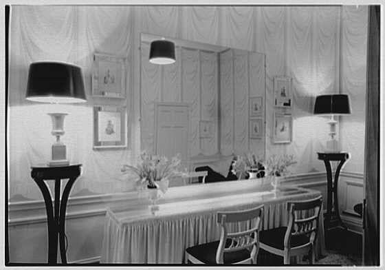 The image depicts a vintage powder room with elegant decor from the year 1946. The space is adorned in monochrome tones, featuring white walls and drapery that create an air of sophistication. On one side, there are two floor lamps standing on either end; each lamp has a dark shade and sits atop small, round tables.

A long vanity stretches across the room with a mirror above it reflecting part of the decor opposite to itself. The countertop is draped in white fabric which adds texture as well as an element of privacy for those using the powder room facilities. Above this area hangs another smaller rectangular mirror framed by what appears to be vintage artwork or photographs.

On top of the vanity, there's a vase with a floral arrangement adding a touch of color and life to the otherwise muted palette of the scene. A couple of chairs are positioned in front of the vanity, likely meant for guests waiting their turn or simply as an inviting seating option.

Overall, this image exudes a sense of classic elegance combined with meticulous attention to detail, characteristic of mid-20th century interior design aesthetics.