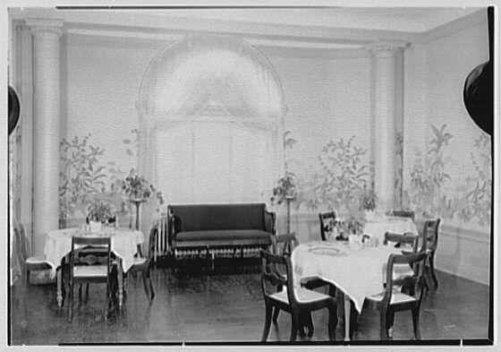 The image is a black and white photograph depicting an elegantly furnished dining area with multiple tables set for a meal. The room has classical architectural elements, such as arched doorways and pilasters on the walls. Each table is draped in a patterned cloth, accompanied by chairs around them. On some of these tables, there are glassware items and possibly dishes or silverware placed neatly. In one corner of the room stands an ornate sofa with dark upholstery that matches the decor's sophisticated tone. The flooring appears to be polished wood, contributing to the overall refined ambiance of the space.