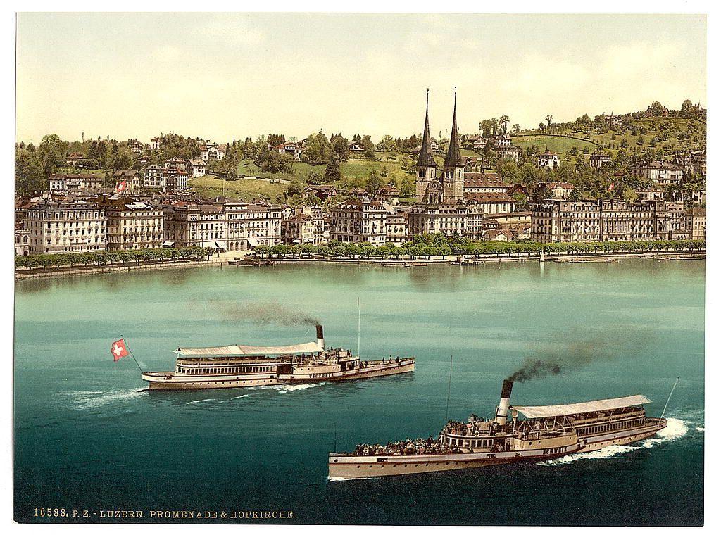 The image depicts a picturesque scene of the Swiss cityscape with historical charm. In the foreground, two steamboats are seen cruising on calm blue waters. The boat closer to the viewer is marked "Promenade & HOFKIRCHE" and bears a prominent red flag bearing a white cross, indicating its association with Switzerland's national symbol.

The backdrop showcases an array of European-style buildings lining the waterfront in Lucerne. They feature traditional architecture reminiscent of the region’s history, including ornate facades and steep roofs characteristic of Alpine towns. Further back, atop rolling green hills, lie two tall spires that appear to be part of a prominent church or cathedral.

The overall impression is one of leisurely travel through an idyllic European setting during the late 19th century, with historical vessels adding character to the scene.