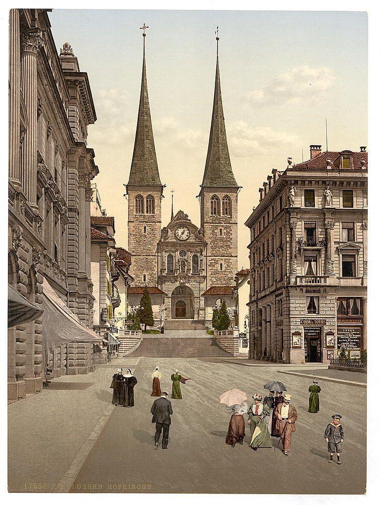 This image depicts a historical scene from Lucerne, Switzerland, captured between approximately 1890 and 1900. The central focus is the St. Peter's Cathedral, recognized by its distinctive clock tower with two spires reaching into the sky. This cathedral serves as an anchor for the composition of buildings surrounding it on both sides.

In the foreground, a bustling street scene unfolds with various individuals dressed in attire indicative of late 19th-century European fashion. Some are walking alone while others form groups, suggesting social interactions or shared destinations. A few people carry umbrellas, possibly to protect against sun rather than rain given the clear sky.

The buildings lining the streets exhibit architectural styles that blend Gothic elements with neoclassical influences, characterized by their ornate facades and detailed stonework. The right side of the image features a row of shops at street level, one prominently displaying "COURT TOURS," hinting at a service-oriented establishment.

The overall atmosphere is lively yet orderly, portraying a moment in time within Lucerne's urban life during that era.