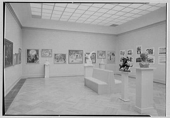 The image shows an interior view of a gallery with black and white artwork displayed on the walls. The gallery has a spacious, open layout with rows of benches along one side for visitors to sit and observe the exhibits. Various paintings and sculptures are placed at different heights throughout the space, creating a dynamic visual display.

On the left wall, several framed pictures hang in a row, depicting various scenes or subjects from art history. The middle part of the gallery has tall pedestals with busts and statues on them, showcasing three-dimensional works prominently for viewers to admire up close.

The right side features another series of artworks displayed at different heights, offering visitors diverse perspectives as they navigate through the space. There is a consistent lighting scheme that highlights each piece individually without causing glare or harsh shadows.

Overall, this image captures an elegant and inviting art gallery environment designed to showcase its collection effectively while encouraging engagement with the artwork from various angles.