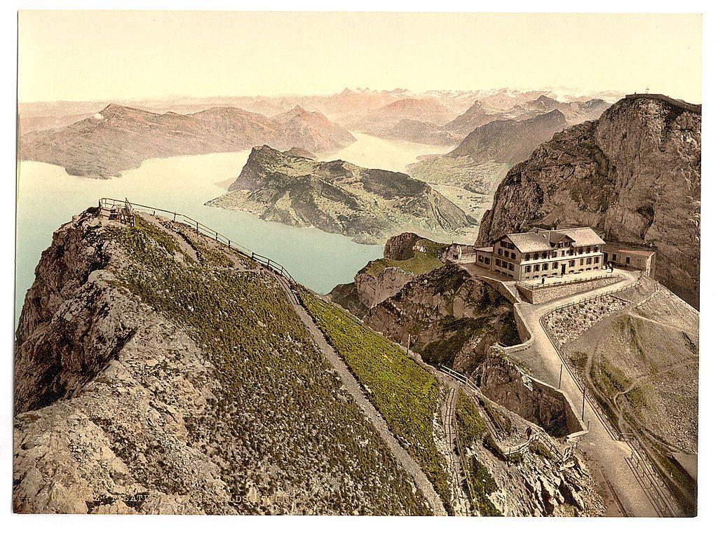 This image depicts a picturesque scene from the mountains overlooking Lake Lucerne in central Switzerland. It appears to be an old photograph, possibly dating back to the late 19th or early 20th century, given its style and coloration.

The landscape is dominated by rugged mountain peaks that rise dramatically against a soft-hued sky. The foreground shows steep rocky terrain with patches of grass and greenery clinging to the slopes, leading up to an observation platform on top. A fenced walkway snakes along one side of this outcrop, providing visitors with panoramic views of the surrounding landscape.

A large building is situated at the summit of the mountain range, likely serving as a hotel or resthouse for travelers. Its architecture blends into its natural surroundings, and it has several wings extending from the main structure. The complex overlooks expansive views of Lake Lucerne below, which mirrors hints of sunlight on its calm waters.

A narrow railway track can be seen snaking down one side of the mountainous terrain towards a tunnel entrance at the base, suggesting that this was once a scenic train route known as "Old Hotel and Esele," leading up to Pilatus. The caption indicates it's from around 1890-1900.

The overall impression is one of historical travel photography capturing the beauty and accessibility of Swiss mountainous regions during an era when such  [...]