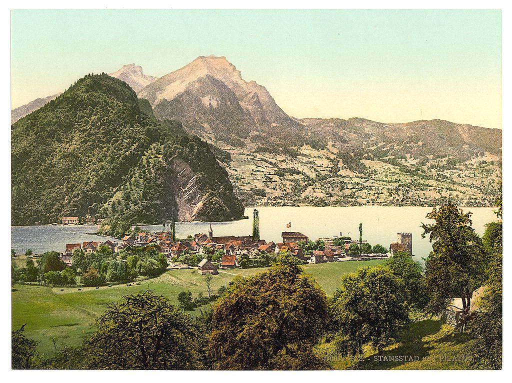 The image depicts a picturesque landscape featuring the town of Stansstad nestled along the shores of Lake Lucerne in Switzerland. The village is set against a backdrop of rolling hills and towering mountains, including Mount Pilatus which looms prominently on one side. The architecture within the settlement suggests historical significance with traditional European buildings clustered together amidst verdant greenery.

The lake itself appears calm, reflecting some blue sky above, while hints of clouds are visible in the distance. A castle or fortified structure stands out near the water's edge, suggesting a strategic location from which to oversee incoming vessels and defend against potential threats.

The overall color palette is warm with rich greens indicating lush vegetation, earthy tones on the rooftops of buildings, and soft pastel blues dominating the skies above Lake Lucerne. The image appears vintage, likely printed during or before World War I based on its style and quality.
