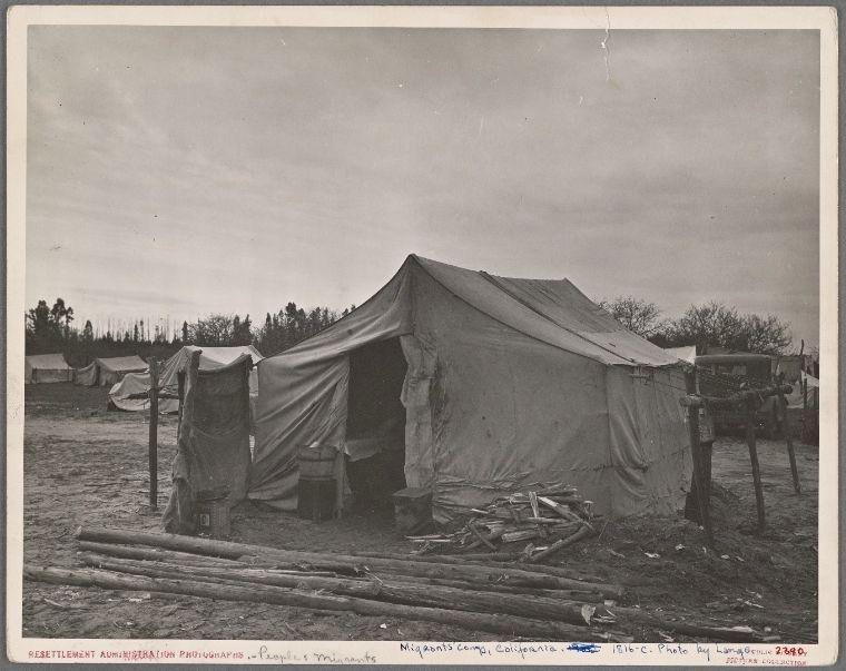 The photograph shows an outdoor setting, specifically what appears to be temporary encampments or makeshift shelters. There is a prominent tent-like structure in the center with various items such as barrels and sticks scattered around it, suggesting that this might have been someone's living quarters during migration or displacement. In the background, there are several more similar tents and structures lined up, indicating an organized campsite for multiple families.

The image appears to be historical, given its black-and-white nature which suggests it was taken in a time when color photography wasn't prevalent or commonly used. The caption at the bottom indicates that this photograph is part of Dorothea Lange's work during her documentation efforts on migrant communities. It specifies "Resettlement Administration Photography—People’s Resettlements" and mentions locations such as Manzanar camp, California.

Dorothea Lange was a renowned photographer known for her documentary photography, particularly focusing on social issues like poverty, immigration, and the impact of war on American society during the early to mid-20th century. The image captured in this photograph is likely one from her extensive work documenting migrant families and their living conditions.

Additional context about Dorothea Lange's photographs can be found through resources such as "Home of a mig [...]