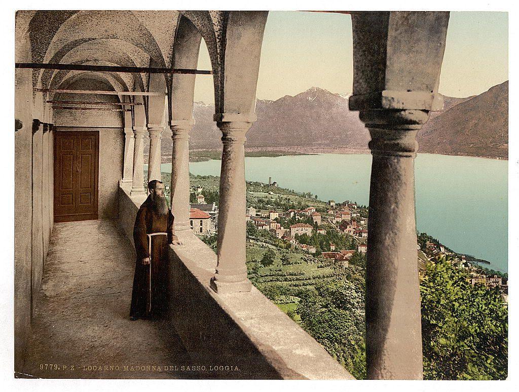 The image depicts a vintage postcard featuring an individual dressed in traditional monastic robes, including a long brown robe and head covering, standing on a stone terrace with arches. The person appears contemplative or is possibly engaged in prayer as they gaze out over the scenic landscape below. They are holding what seems to be a book or religious text.

The backdrop reveals a picturesque town nestled alongside a lake surrounded by lush greenery and mountains under a clear sky, suggesting a serene and remote location. This setting indicates that it might represent Loggia, located in Locarno near Madonna del Sasso in Switzerland during the late 19th century to early 20th-century timeframe.

The image exudes an atmosphere of tranquility, spirituality, and historical charm, showcasing the beauty of Swiss landscapes from a bygone era.