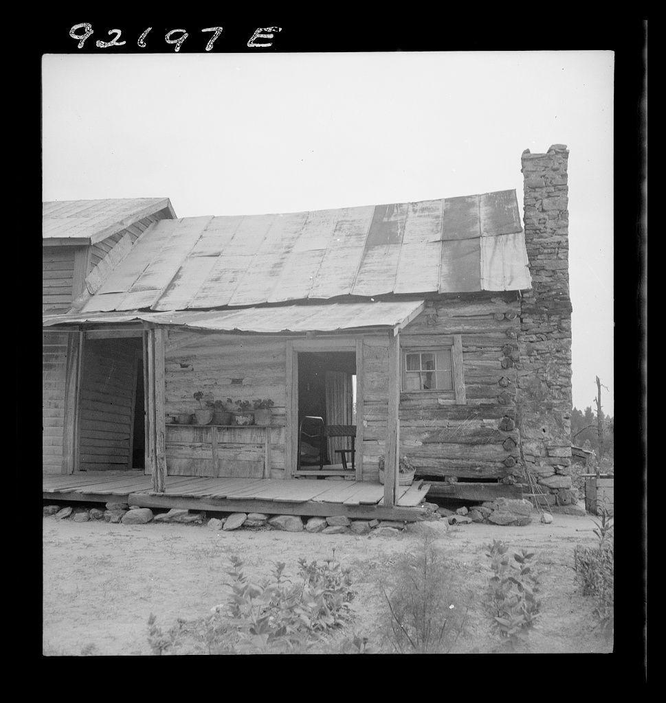 The image depicts a rustic wooden cabin with weathered siding and an uneven roof made of corrugated metal. The structure shows signs of age, such as exposed beams and peeling paint on the exterior walls. To the right side, there is a stone chimney that stands tall above the roofline. On the porch area, various items like pots with plants are placed along the front steps leading into an open doorway.
Behind this cabin lies another structure made from logs, which appears to be even older and more worn than the first one. The ground around both cabins shows patches of grass and dirt, suggesting a rural or semi-rural setting. In the foreground on the right side, there is some vegetation that could possibly be part of a garden.
The image has a handwritten date "92197 E" at the top left corner, indicating it may have been taken in 1921 based on its composition and quality. This black-and-white photograph captures an essence reminiscent of early to mid-20th-century rural life, possibly from North Carolina as mentioned in the caption.