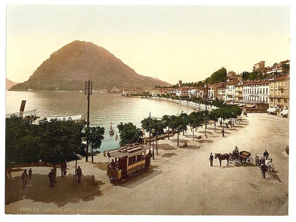 The image is a sepia-toned photograph depicting a scenic town landscape, likely taken around the late 19th or early 20th century. In the foreground, there's a cobblestone street with several individuals walking and three horse-drawn carriages: one appears to be an old-fashioned tram-like vehicle painted yellow with black details, while two others are smaller wagon-type vehicles attached to horses.

On either side of this central thoroughfare is a row of European-style buildings with uniform facades featuring shuttered windows. The architecture suggests the location could be in Europe or possibly Switzerland, as indicated by the text at the bottom referencing Lugano and San Salvatore. A prominent mountain looms large on one side across what appears to be a calm lake.

On this lakeside promenade are people strolling along with trees lining both sides, suggesting an attractive riverside town setting. The overall atmosphere is peaceful, reminiscent of leisurely late 19th-century European life.