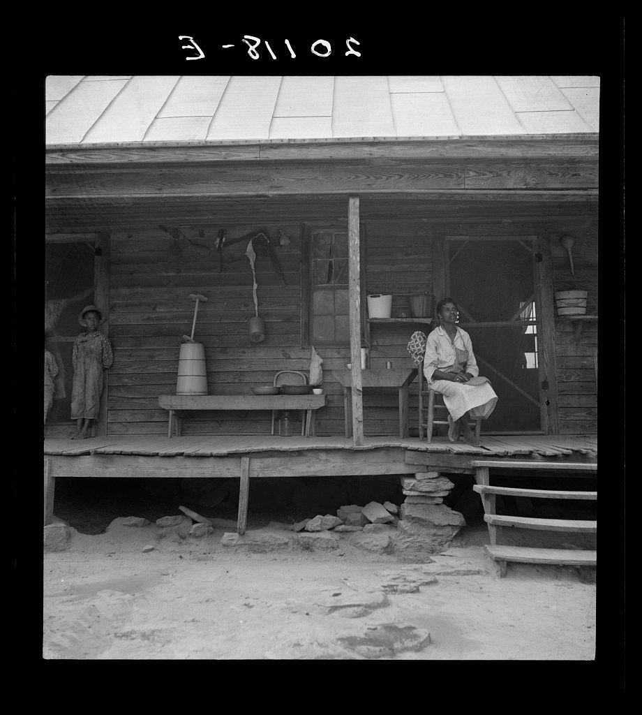 A black and white photograph shows an older woman seated on the porch of a wooden house, possibly from early to mid-20th century judging by her attire. She is engaged with something in her hands, potentially reading or handling some papers. The porch displays various household items such as pots, pans, utensils, and cleaning tools hung up for convenience and perhaps drying after use.

A young child stands on the left side of the frame, dressed in simple clothing that suggests a rural setting. Both figures exhibit a calm demeanor typical of the time period's photographs which often captured everyday life with dignity and authenticity. The environment appears modest but functional, indicative of rural living conditions during that era.
