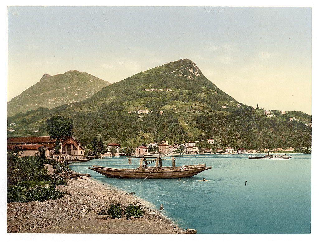 This image depicts a picturesque lakeside scene with mountains in the background and several buildings along its shore. In the foreground, there is calm blue water where traditional wooden boats are docked or floating gently. One of these boats has two individuals aboard it, possibly engaged in some form of laborious activity such as fishing or transport.

The midground features more vessels on the lake, with one boat closer to a small hillside village nestled among terraced gardens and vineyards that rise up the slopes leading to the mountainside settlements. The lush greenery indicates this might be during late spring or summer months when crops are well established but not yet fully ripened.

On land near the water's edge, there is an old-style building with a gabled roof adjacent to another structure with red roofing and visible structural details suggesting historical significance. This setting suggests that the photograph was taken at Cassarate on Lake Lugano in Switzerland during approximately 1890-1900.

The overall atmosphere of this scene exudes tranquility, capturing everyday life within a picturesque Swiss landscape from over a century ago.