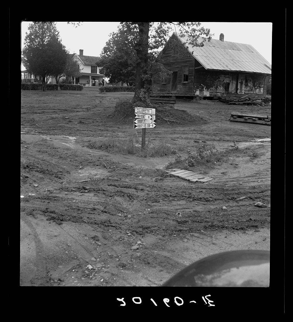 The image depicts an outdoor rural scene with a mixture of natural and man-made elements. At the center, there's a dirt patch that appears to be part of a yard or field, showing signs of recent activity as indicated by tire tracks embedded in it.

Three wooden directional signposts are prominently positioned on a pole at one end of this area. The topmost sign reads "FILLED WITH," followed by two smaller directions indicating turns and distances towards various places: "PROVIDING SHOFLY 195-4" to the right, pointing presumably northwestward, with another arrow suggesting a route labeled as such.

Surrounding these signs are modest structures; on one side stands an old-fashioned house featuring what looks like a porch supported by columns. It's white and has visible architectural details hinting at its age or construction style typical of rural North Carolina during the early 20th century, judging from stylistic elements present in similar period photographs.

Adjacent to this is another wooden structure with a slanted roof and a front facing an open field. The simplicity of these buildings underscores their utilitarian purpose, likely serving as homes for residents or perhaps storage for farm-related equipment given its proximity to the dirt patch which could be indicative of agricultural activities.

The overall atmosphere evokes one from past times in rural life, possibl [...]