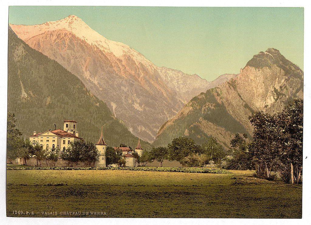 The image depicts a picturesque landscape featuring the Château de Werra located in Valais, Switzerland. This early 20th-century photograph captures a vast green field leading up to an imposing chateau surrounded by fortified walls and towers. The castle is painted in light beige with dark red roofing that contrasts beautifully against its natural surroundings.

In the background, majestic mountains rise dramatically from the valley floor, their peaks partially covered with snow suggesting they could be part of the Pennine Alps or related mountain ranges nearby. The coloration has a vintage hue typical of chromolithographs produced during this era, enhancing the historical charm and appeal of this Swiss landmark.