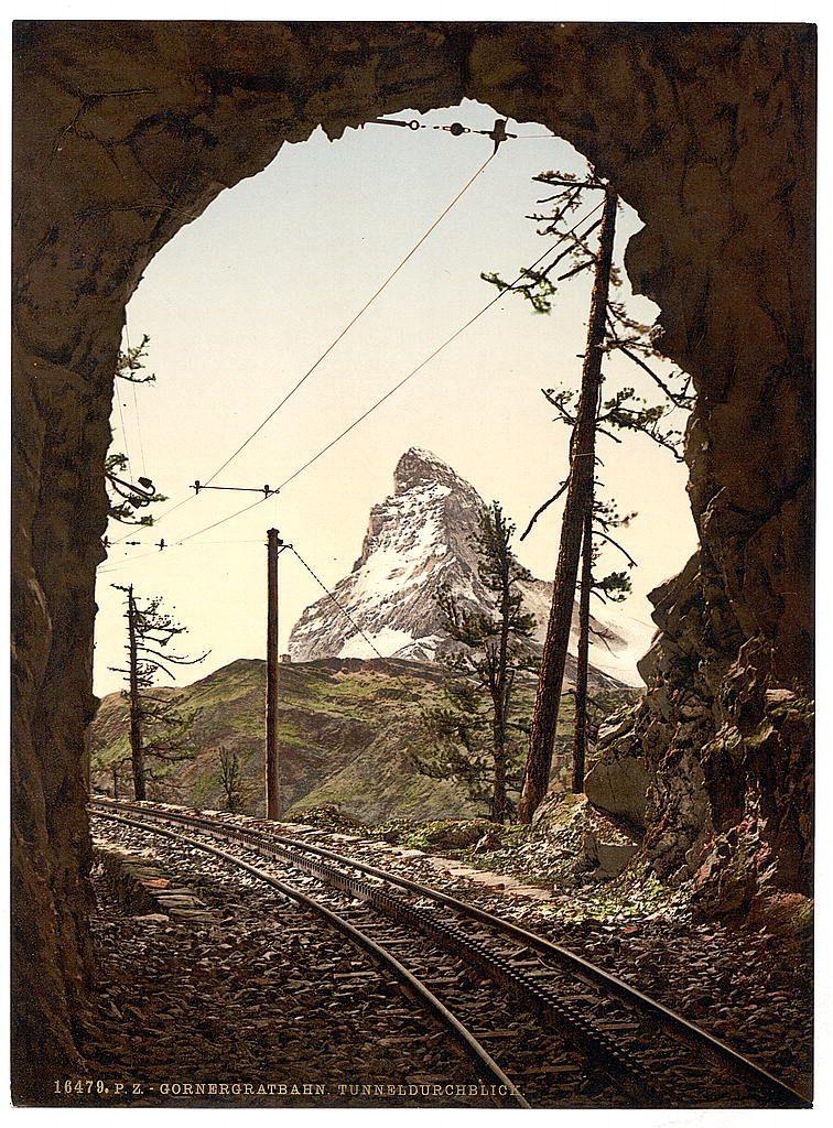 The image shows a scenic view through an arched tunnel, revealing railway tracks leading into the distance. The focus is on a majestic mountain with its peak covered in snow and partially shrouded by clouds or mist at sunrise or sunset, suggesting either early morning or late afternoon light conditions. Towering above are coniferous trees indicative of high-altitude forestry typical to such regions. The surrounding landscape features gentle slopes adorned with patches of greenery, hinting at the season being possibly spring or summer due to visible foliage.

The railway tracks belong to a historic cogwheel rail system known as Gornergratbahn in Switzerland's Valais region within the Alps. This mode of transportation was pivotal for tourism and connectivity during its operational years between approximately 1890-1900, catering to tourists eager to explore this part of Europe from an elevated vantage point.

The photograph appears vintage with a sepia tone common to early postcards or colorized prints of that era. Details such as the railway signaling infrastructure and overhead lines emphasize both its utilitarian purpose and the aesthetic allure it offered to travelers of the time, providing them picturesque views like this iconic one featuring the Matterhorn from Gornergratbahn tunnel vantage point.

The caption "18479. P-Z - GORNERGRATBAHN, TUNNELBILD" suggests that thi [...]