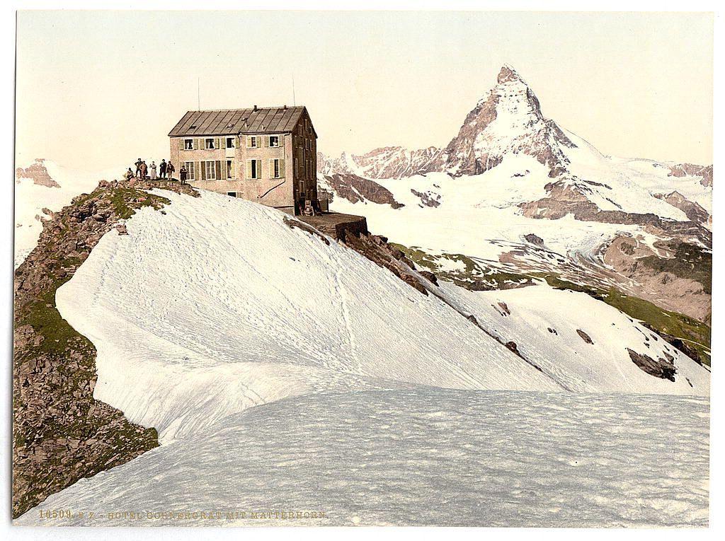 The image depicts a historic scene, likely from the late 19th or early 20th century. It features a large stone building situated on an elevated mountainous terrain covered with snow and patches of exposed rock. The architecture suggests it is either a hotel or lodge, given its multiple windows and pitched roof typical for such establishments in high-altitude environments to cope with harsh weather conditions.

In the foreground, we observe a group of people standing near what appears to be a small wooden platform or staircase leading up to the building's entrance. Their attire indicates they are dressed warmly, likely due to the cold climate suggested by the snow-covered landscape around them. They seem engaged in conversation and appear relaxed despite being at such an elevation.

The background is dominated by a prominent mountain peak with its distinctively pointed summit, indicative of alpine topography like that found in Switzerland's Alps or possibly the Dolomites further north on Italy-Slovenia border region. The snow cover indicates it could be late spring to early summer when temperatures are still below freezing at higher altitudes.

The overall composition and details suggest this image is a historical postcard, capturing an era where such mountaintop accommodations were attractions for tourists seeking adventure or relaxation in picturesque natural settings.