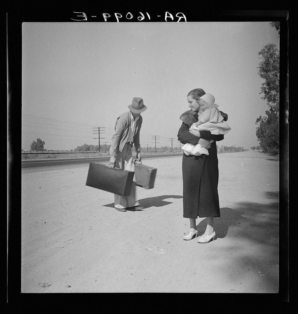 The image is a black and white photograph depicting three individuals on what appears to be an empty road. On the left, there's a man wearing a suit with a hat, looking down as he lifts two large suitcases one in each hand. In the center of the frame stands a woman holding a baby close to her chest; she wears a long skirt and high-heeled shoes while clutching what looks like a fur-trimmed coat or wrap around herself. To her right is another child, presumably hers as well, who sits on her lap wearing a cap. The background shows an expansive road leading towards the horizon with minimal structures visible beyond it - telephone poles and sparse vegetation under clear skies suggest this may be in California's rural areas during 1935 when such families often traveled via highway for seasonal farm work or due to economic hardships caused by Great Depression conditions prevalent across America.