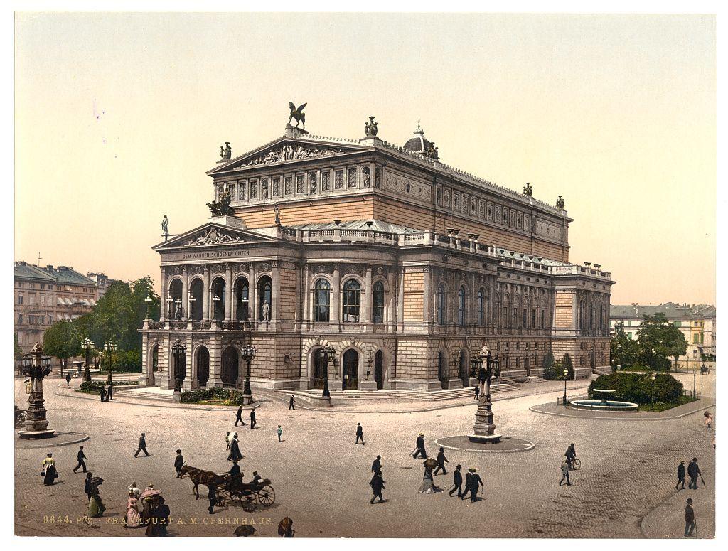 The image displays a historic postcard featuring an opera house building in the center, identified as "Opera House" located at Frankfurt am Main. The architecture of the structure is grand and classical with ornate detailing on its facade, including sculptures above the entrance and decorative cornices along the top edge.

In front of the Opera House, there's a bustling street scene depicting late 19th or early 20th-century life. People are seen walking in various directions, engaging in conversation, and going about their daily activities. A horse-drawn carriage with two people is prominently visible on one side, suggesting transportation modes from that era.

The surrounding area includes other buildings of different architectural styles, indicating an urban setting. The sky appears clear, hinting at a pleasant day during the time period represented by the image's context clues.

Notable features include vintage clothing styles worn by individuals and the overall sepia-toned color scheme typical for such historical postcards from that era. There are no modern elements or indications of contemporary times within this scene.