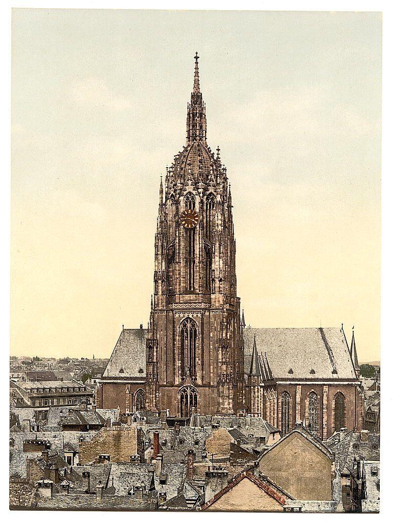 The image depicts a grand Gothic cathedral, characterized by its intricate stone architecture and towering spires. The structure features large arched windows with tracery, flying buttresses for support, and an elaborate facade adorned with statues and decorative elements. At the pinnacle of the church stands a tall steeple crowned with a cross, adding to the vertical emphasis typical of Gothic design.

The cathedral is surrounded by densely packed rooftops of surrounding buildings, suggesting that this image captures it within or near a bustling urban area. The photograph appears aged, as indicated by its sepia-toned coloring and visible wear on the paper. This historical document might have been created between approximately 1890 to around 1900.

The landmark is identified as Frankfurt Cathedral (Frankfurt am Main), Germany based on archival information from Views of Germany[^2^]. The image serves both a historic record and an artistic portrayal, showcasing architectural beauty while also providing context about the urban environment in which such grand structures stood.