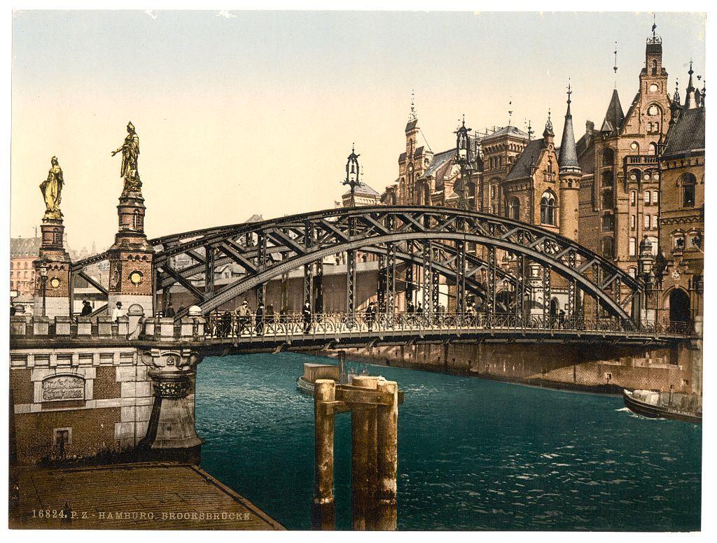 The image depicts a historic bridge over a river in Hamburg, Germany, with the caption indicating it is from Brooksbridge (Brooksbrooke). The photograph appears to be vintage, possibly dating back to around 1890-1900. It shows an ornate steel truss arch bridge spanning across calm waters beneath. On either side of the riverbank are statues on pedestals and a stone embankment with architectural features typical of late 19th-century European urban design, including pointed towers and decorative stonework.

In the background stands an elaborate building complex featuring Gothic Revival architecture characterized by steeply pitched roofs, tall spires, arched windows, and ornate façades. The sky is overcast or colored with a sepia tone typical of old photographs, enhancing its historical feel. Boats are visible on the river below, suggesting active waterway use at that time.

The overall scene captures an early urban landscape in Hamburg during a period when such infrastructure was being developed and celebrated as modern marvels by contemporary society.