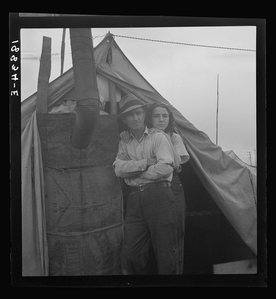 The image shows two individuals, likely middle-aged men and a younger woman or young adult female. They are posing for the camera with their arms around each other in front of what appears to be makeshift canvas tents or temporary structures commonly used by migrant laborers during agricultural seasons. The man on the left wears a wide-brimmed hat and has his hand resting over one shoulder, while the person beside him is slightly leaning into the frame.

The black-and-white photograph seems historical, possibly taken in California due to its mention of Near Strathmore, CA along with Wyoming and Missouri origins mentioned alongside "eight years in California working in lemons." The individuals are dressed casually for outdoor work; one wears a long-sleeved shirt and trousers while the other is also clad similarly.

The overall setting suggests a tough lifestyle often associated with migrant workers during agricultural seasons.