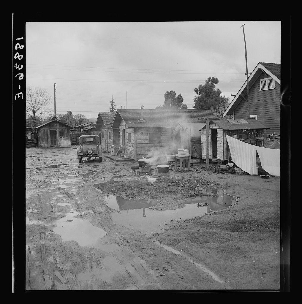 The image is a black and white photograph depicting what appears to be an old-fashioned, impoverished living area. Several small wooden structures with weathered exteriors line the scene, including huts and a larger house-like building emitting smoke from its chimney, suggesting that it may contain a wood stove or similar heating mechanism inside. The ground shows evidence of recent rain as puddles have formed on the dirt path leading through what seems to be an alleyway between these structures.

A dilapidated truck is parked in front of one of the wooden buildings, indicating some form of transportation for the inhabitants. There are various items scattered around outside: a bucket, clothes hanging out to dry, and other miscellaneous objects that suggest daily living activities. The environment looks unkempt with signs of disrepair on both man-made structures and natural elements like grasses.

In summary, this image portrays a modest dwelling area in possibly rural or semi-rural California during what could be the early 20th century based on architectural styles and conditions presented.