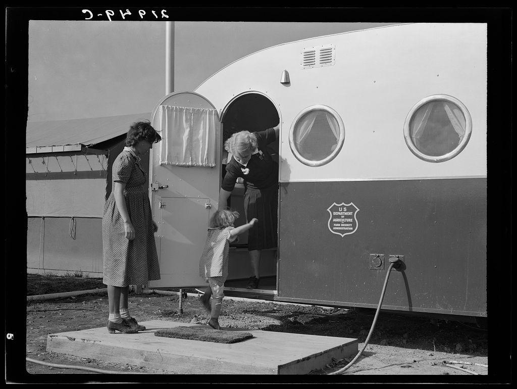 The image depicts a moment in time involving three individuals, likely from an earlier era given the style of clothing and black-and-white quality. On one side stands a woman dressed in a patterned dress with rolled-up sleeves, wearing dark shoes adorned with garter belts, suggesting practical footwear suitable for outdoor activities or travel. She appears to be supervising or waiting as she looks towards another figure.

In contrast, this second person is not fully visible; we can only see their torso and arms through the doorway of a trailer marked "U.S. Department Agriculture Agricultural Extension Service." The individual seems engaged in an activity within the trailer's interior space. Another key subject stands outside on what appears to be a makeshift platform or step.

The exterior features include circular windows with distinctive framing, likely for ventilation purposes, and an open entrance allowing us glimpse into another room. There is also a hose connected via a fitting protruding from one side of the vehicle, indicating utility access possibly related to sanitation needs like washing facilities in this particular setting.

Overall, the composition of elements suggests a temporary settlement or camp environment where services such as medical examinations are provided by extension service representatives within trailers designated for these purposes. The imag [...]