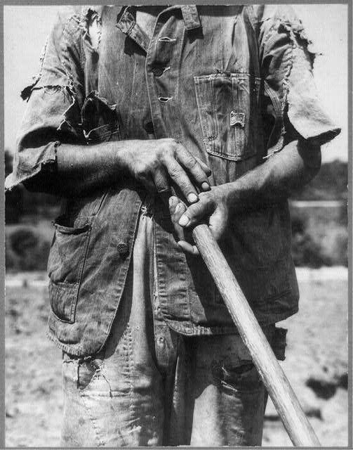 A black and white photograph of an individual dressed in a heavily worn, denim overalls suit. The person's hands are placed together across their stomach as they grip the handle of what appears to be a farming tool or implement, likely indicative of labor-intensive work such as farming. Visible signs of distress or fatigue can be seen on the face through the eyes and eyebrows. No background elements provide context for location but seems open-air given lighting conditions. The image is labeled with an alphanumeric code beneath it which suggests some form of cataloguing system was used to identify this particular photograph.