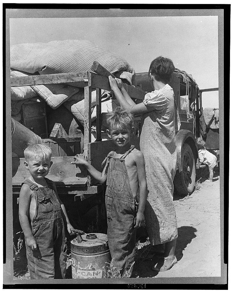 This black and white photograph captures an impoverished family of nine, including a sick four-month-old baby. They are traveling down New Mexico highway on foot as refugees from Iowa due to the Great Depression in 1932 because of their father's ill health. The man was once an auto mechanic laborer who is also tuberculosis-affected and unable to work anymore.

The family has been living off relief, with some time spent in Arizona before being refused entry onto any more relief roles on a return attempt to Iowa due to the strain they could be expected to impose upon those states' resources. The caption notes that they have no money at all left as their belongings and trailer are about to be sold for food.

The image evokes an era of hardship, with one girl holding up wooden planks in front of a barrel labeled "canola oil," suggesting the family is seeking any available means of sustenance during this time. The visual representation offers insight into the struggles faced by families displaced and struggling due to economic downturns or natural disasters like tuberculosis outbreaks.