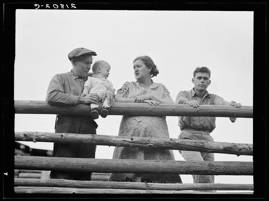 The image is a black and white photograph showing four individuals standing behind a wooden railing. From left to right, there's an older man wearing a hat, holding a baby; next to him is a woman in a dress with her mouth open as if speaking or singing; beside the woman stands another young adult male looking away from the camera with his hands on the rail; and lastly, there's a younger individual, possibly late teens or early twenties. The clothing suggests this could be a historical photo, potentially representing rural life of that era in Idaho.