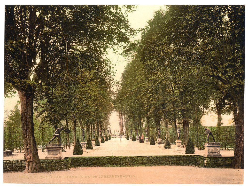 The image depicts a formal garden pathway lined with evenly trimmed hedges and mature trees. There are several statues placed on pedestals along the path, which appear to be classical in style. The perspective is from within the garden looking down the length of the path towards what seems like an entrance or focal point at the far end. Overhead, branches hang over some parts of the pathway, suggesting a shaded environment with dappled sunlight filtering through. In the distance, two individuals can be faintly seen walking along the path. The overall atmosphere is serene and reminiscent of classical European gardens found in historic estates or public parks from around that era.

The image appears to have been printed using a chromolithographic process typical for postcards or prints during late 19th to early 20th century, with colors faded over time but still discernible. The bottom text indicates the location as "Sommertheaterz" in Herrenhausen, Hanover, Germany and suggests it was taken between approximately 1890 and 1900 based on historical context of such prints.

This photograph is part of a collection that includes other images related to German views during this period.
