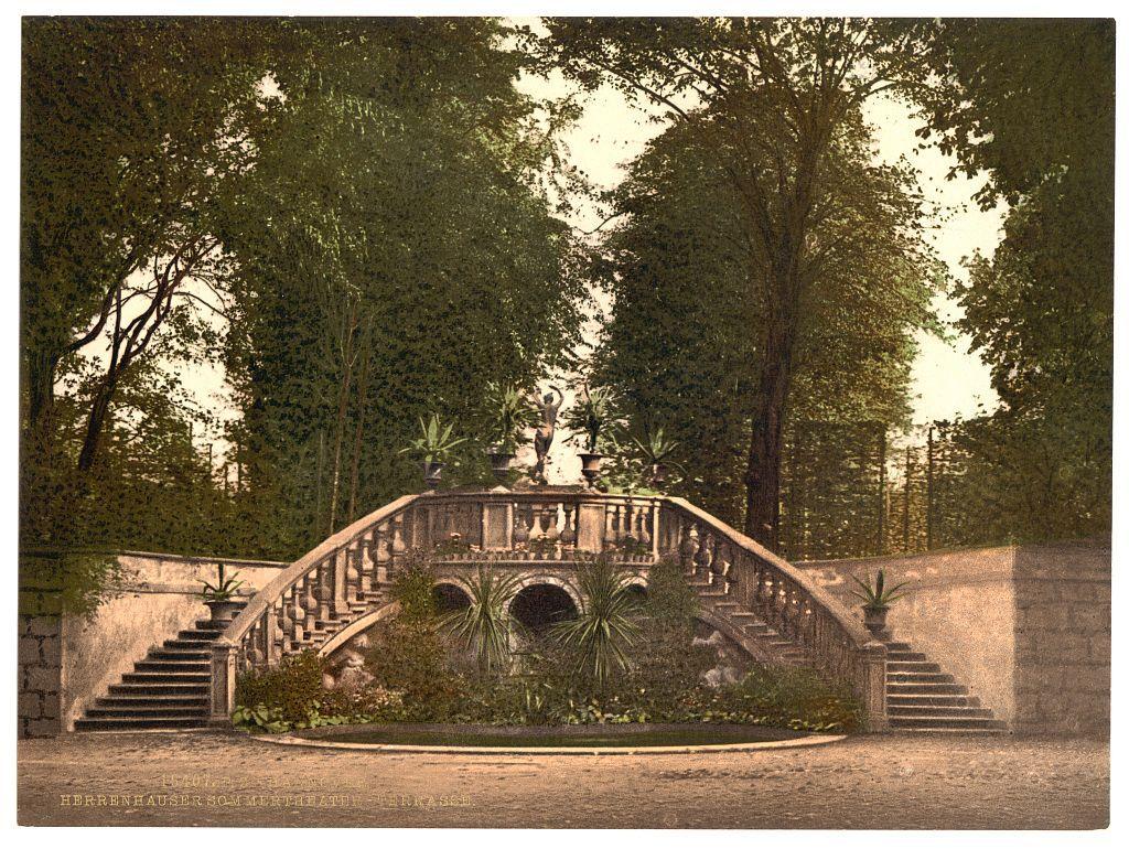 The image depicts an ornate staircase leading up to a bridge in what appears to be a lush garden setting. The stairs are constructed with stone, featuring detailed carvings and decorative elements such as urns at the railings' ends. Atop this bridge is another smaller archway or tunnel-like structure adorned with additional greenery. Lush foliage surrounds the staircase and extends into the background where several trees provide a dense canopy of leaves. The scene suggests an early 20th-century European garden, possibly part of Herrenhausen Gardens in Hanover, Germany, as indicated by the text at the bottom left corner which also includes German script likely referencing "Herrenhaus."
