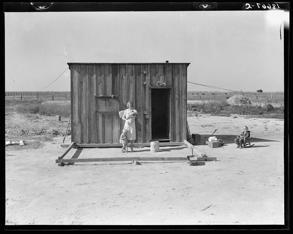 The image depicts a black-and-white photograph of an outdoor rural setting, showing two individuals and various items around them. In the center stands a small wooden shack with visible vertical planks forming its walls, a single door on one side, and a window above it to let light in. The roof is made up of what appears to be corrugated metal sheets.

In front of the shack, there's an adult female standing with her arms outstretched holding two young children - possibly her own kids or relatives being cared for by someone else. She has short hair and wears a dress that seems suitable for warm weather conditions; she is barefoot on what looks like dusty ground indicating arid landscape.

To the right side of this image, there's an older male seated comfortably in a wooden chair made from tree branches with attached slats forming backrests. He holds his hands together or perhaps has something small between them as he gazes off to one side thoughtfully, suggesting contemplation or waiting for someone.

The surrounding environment is open and spacious with no visible structures other than the shack which might be their home; it seems quite remote from urban areas considering its isolated position within a vast expanse of land. The ground around appears dry without any vegetation indicating either seasonal drought conditions or typical climate where such landscapes are common [...]