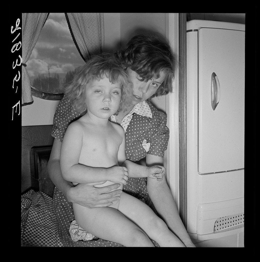 The image is a black and white photograph depicting two individuals, likely mother and child. The adult appears to be examining or comforting the young person seated in her lap. They are inside what seems like an RV or trailer setting, indicated by the curtains on one side of them and part of an appliance that could be a refrigerator visible beside them.

The adults's attire suggests early mid-20th-century fashion; she wears a polka-dotted dress with a white collar while holding something in her other hand. The child has curly hair and is not wearing any top, revealing their bare chest, which may indicate they were undressed for the examination or due to discomfort.

The setting conveys an intimate domestic scene, possibly related to health care within the context of trailer living during that era as suggested by associated captions mentioning "Doctor examining children in trailer clinic" and a reference to FSA (Farm Security Administration) mobile camps.