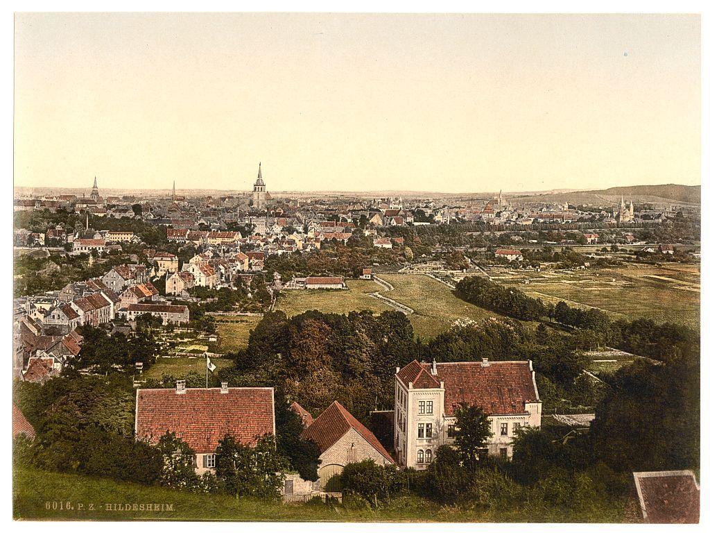 The image presents a scenic panoramic view of the town Hildesheim in Hanover, Germany. It is an old photograph with sepia tones and hand-colored areas that highlight certain features such as roofs, trees, and fields.

In the foreground to midground are clusters of buildings indicative of residential structures; their architecture suggests European style housing from a historical period—likely late 19th or early 20th century. Prominent in this area is what appears to be an estate with well-kept gardens, surrounded by red-tiled roofs and greenery.

The background reveals the expanse of Hildesheim's urban landscape extending into the distance where more buildings are visible along with towers that could signify churches or other significant structures common in European towns. The overall impression is one of a thriving community nestled within a hilly terrain, likely on higher ground given the perspective from which this photograph was taken.

The sky above has been rendered lightly and lacks detail, focusing attention below where architectural interest lies—the buildings stand out against an almost pastel-colored skyline with hints of blue near the horizon. Notable is the absence of modern elements like cars or contemporary infrastructure; it’s a view that captures Hildesheim in what seems to be its historical state.

This photograph carries significant cultural and histor [...]