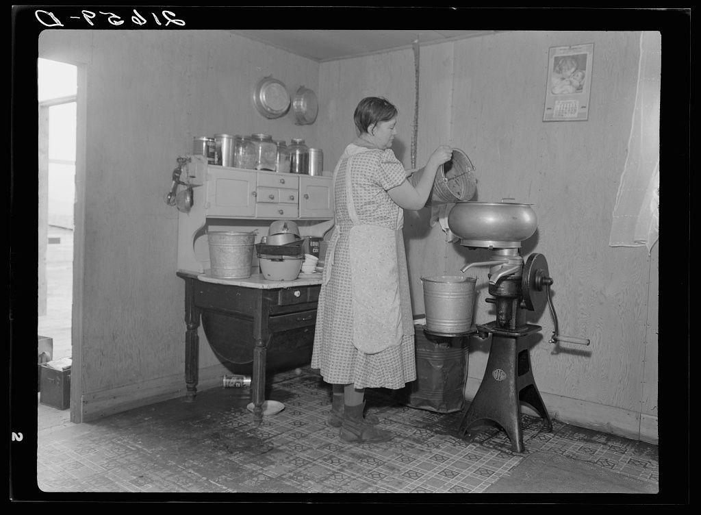 The image shows a black and white photograph of an individual in what appears to be a domestic setting, engaging with kitchen appliances. The person is wearing a patterned dress or apron over lighter-colored clothing, suggesting they may have been involved in food preparation or cooking activities.

In the background, various items such as jars on a shelf, a calendar hanging on the wall, and containers likely used for storing ingredients are visible. There's also what appears to be a small kitchen table with objects placed upon it. To the right side of the image is an old-fashioned manual grinder or miller that has been in use.

The environment suggests this could be from an earlier time period when such appliances were common, indicating perhaps rural life conditions where homes had basic amenities for food preparation but no modern electrical conveniences like a blender or mixer yet existed. This setting may evoke themes of historical living standards and domestic practices during the early to mid-20th century in America.

The photograph is labeled with "Dorothea Lange," which could indicate that it was taken by Dorothea Lange, an American photographer known for her work documenting social issues such as poverty, migration, rural life, class conflict, and industrialization. The reference number 'C-10625' might be related to a collection or archive where this photograph  [...]