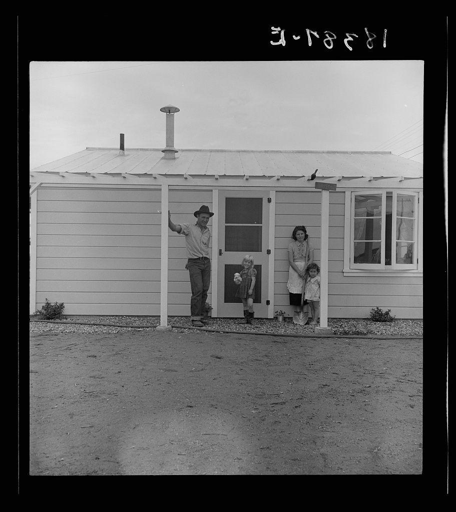 The image shows a black and white photograph of an older man, woman, and two children standing in front of a modest single-story house. The man is wearing a hat and seems to be waving or gesturing with his right hand raised above his head as he stands on the porch area which has sliding glass doors partially open. He appears to be welcoming someone into his home.

The woman is holding one child's hand, while the other child holds onto her arm. The older man in the picture looks cheerful and seems proud of their new housing situation. Both adults wear casual clothing appropriate for a rural setting; the man wears rolled-up jeans and shoes suitable for farming work while the women are dressed modestly.

The children appear to be girls based on their attire, one wearing overalls with a patterned top, and another in lighter-colored clothes possibly made of cotton or linen. They both look content but perhaps not as excited about being photographed compared to the adults' enthusiasm.

There's vegetation around the house which appears to have been recently tended; small bushes are visible beside the porch on either side. The ground is unpaved dirt with a slightly rough texture, indicating it might be outside of an urban area in California where this was likely taken during the time when Dorothea Lange documented migrant workers' living conditions.

The photograph captures emotio [...]