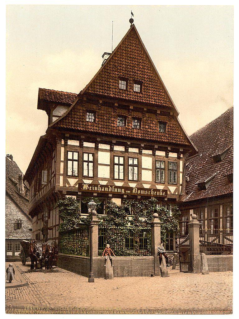 The image depicts a historical European building with architectural features typical of the late 19th or early 20th century. It is constructed in the traditional half-timber style, characterized by wooden beams exposed on its upper floors and white plaster infill between these timber frames. The roof appears to be steeply pitched, covered in red tiles that are common in Northern European architecture.

The building has multiple levels with a prominent triangular gable at one corner displaying ornate signage reading "Wienhandlung um Domscheine," which suggests it was used for trading religious objects or as an inn. The facade is adorned with climbing ivy, adding to its aged charm and hinting that the structure might be of considerable age.

In front of the building stands a woman dressed in period-appropriate attire: she wears a long dress paired with boots, indicative of late 19th-century fashion for women. She appears contemplative or is perhaps waiting for someone. Near her are two stone pillars supporting what seems to be an ornamental lamp post; another pillar and railing can be seen further back.

There's also a horse-drawn carriage parked beside the building, reinforcing the historical context of the photograph. Another individual sits on steps leading up to the doorway, adding life to this otherwise still scene.

The overall setting appears calm with no apparent hu [...]