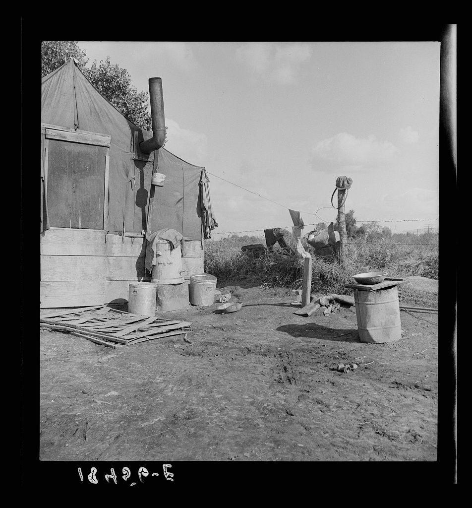 The image is a black and white photograph depicting an outdoor scene, likely from the mid-20th century based on its style. The setting appears to be rural with sparse vegetation in the background under a clear sky. Prominently featured are makeshift wooden structures, possibly temporary housing for migrant workers or agricultural laborers.

To the left is a large canvas tent supported by poles, equipped with various items attached externally such as a clothesline with garments and other miscellaneous objects like buckets, pails, and what looks to be cooking utensils on stands. Adjacent to it are barrels of different sizes suggesting storage for water or supplies.

In front of these tents, there is an array of wooden planks scattered haphazardly across the ground. To the right side, a tall chimney pipe extends vertically from one structure towards the sky, indicating some form of heating system within this makeshift shelter.

The overall environment conveys simplicity and resourcefulness, with signs of wear evident on both structures and materials suggesting prolonged use or lack of maintenance. The absence of electrical appliances suggests limited amenities.

There are also two buckets in front of what appears to be a larger storage vessel; one bucket is overturned beside it. A small bowl sits atop the top of this container which could suggest food preparation taking plac [...]