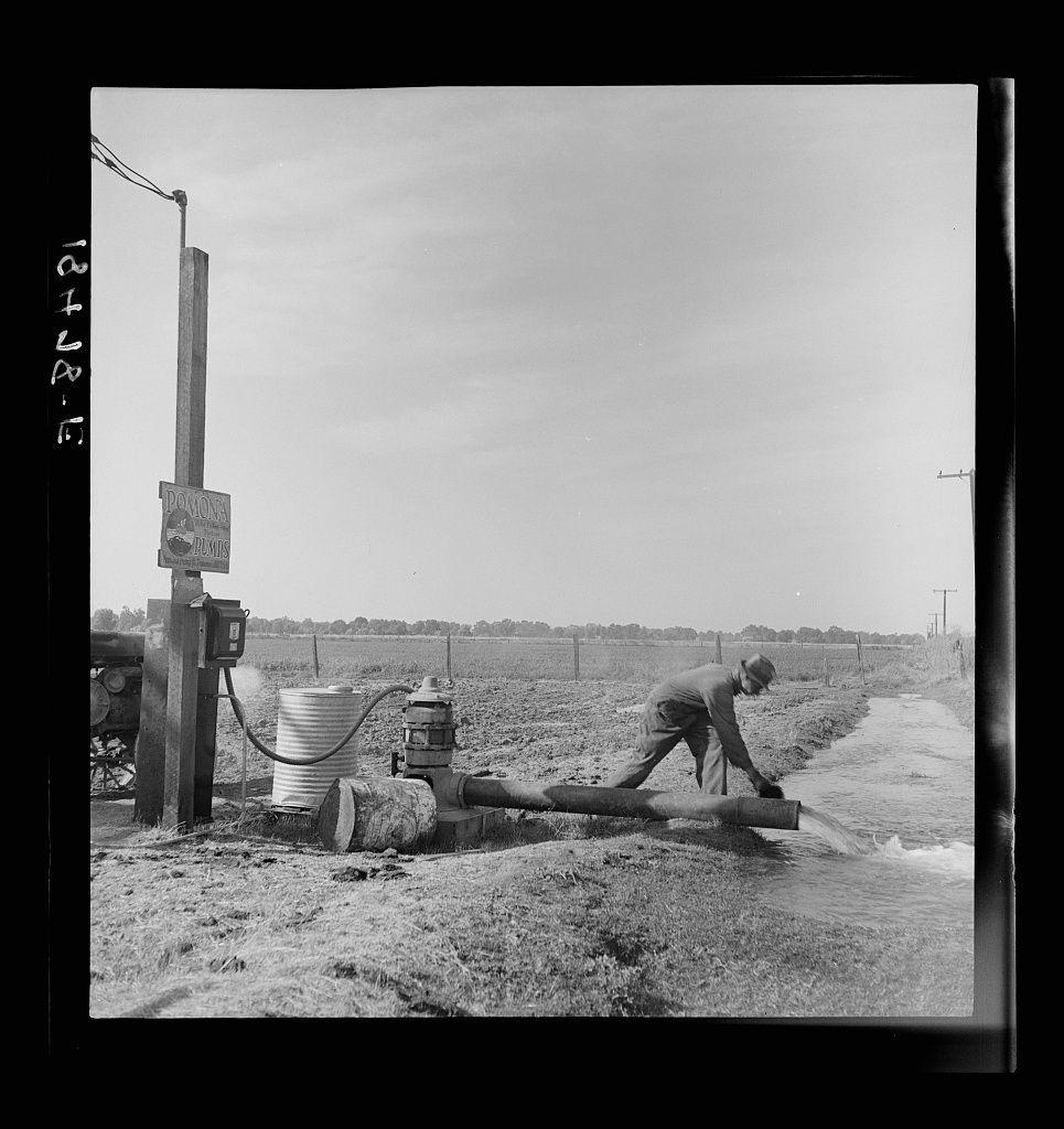 The image shows a monochromatic scene of what appears to be an irrigation pump located on the edge of an agricultural field. A person is depicted in mid-action, actively engaged with the equipment - seemingly operating or maintaining it as water flows from pipes into the surrounding area which seems like a small canal for distribution.

In the background, we see vast open fields stretching towards distant treelines under what could be interpreted as a clear sky based on the brightness and lack of visible clouds. The field is marked by furrows typical of plowed land ready or post-sowing conditions in farming practices.

The equipment includes a large cylindrical tank connected to several pipes with valves, which suggests its function is likely related to water management for irrigation purposes. A wooden pole supports what looks like an electrical box and possibly power lines above it - indicative that this operation relies on electricity rather than manual labor alone. 

Additional structures include simple utility poles in the background, reinforcing a rural or semi-rural setting where such agricultural practices are common.

There is visible text on a signpost; however, due to image resolution constraints, its exact content isn't discernible here but it contributes contextually as potentially related information regarding irrigation control or operation.