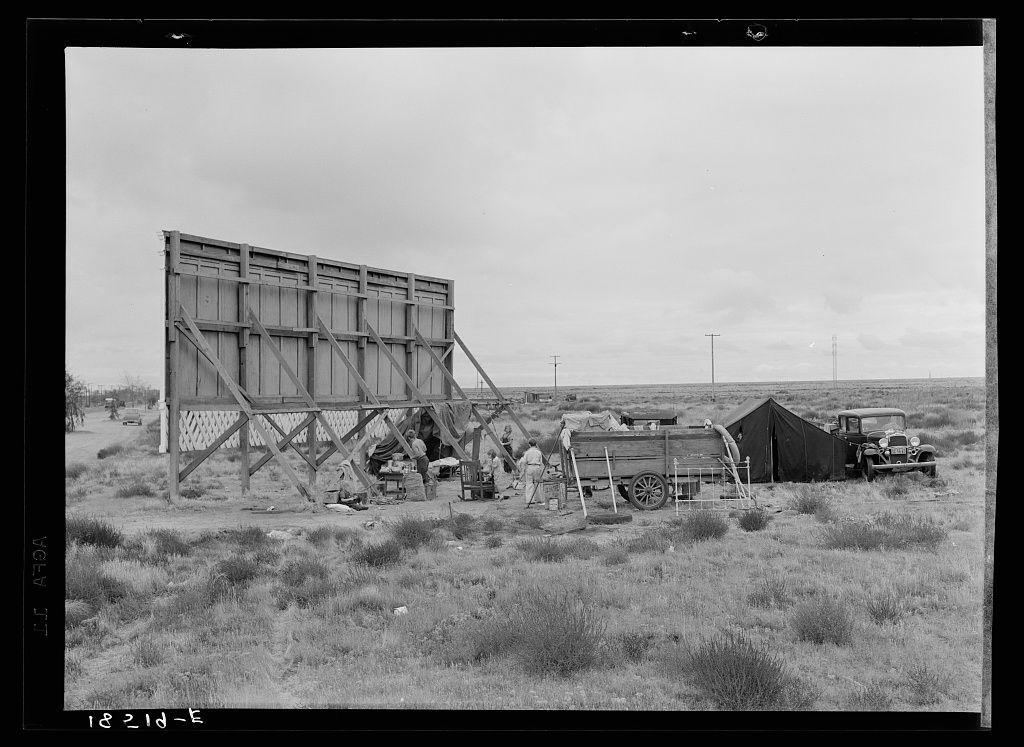 This black and white photograph captures a scene of rural life, possibly during the Great Depression era. In the foreground, three individuals are seated on wooden stools under an unfinished structure that appears to be in various stages of construction or disrepair. The figures seem engaged in conversation with one another.

The middle ground is occupied by two automobiles: a truck and what looks like a sedan, parked beside each other. Next to these vehicles stands a large, makeshift billboard sign propped up on wooden beams, which suggests the area might be used for advertising or public notices during gatherings.

In the background, a simple tent pitched between poles indicates that this gathering is likely temporary accommodation rather than permanent housing. The landscape around them consists of dry grass and sparse vegetation, hinting at an arid environment with limited resources.

The overall composition conveys a sense of resilience in the face of challenging circumstances, as these individuals have come together to create community amidst their nomadic lifestyle on what could be considered America's "Main Street."