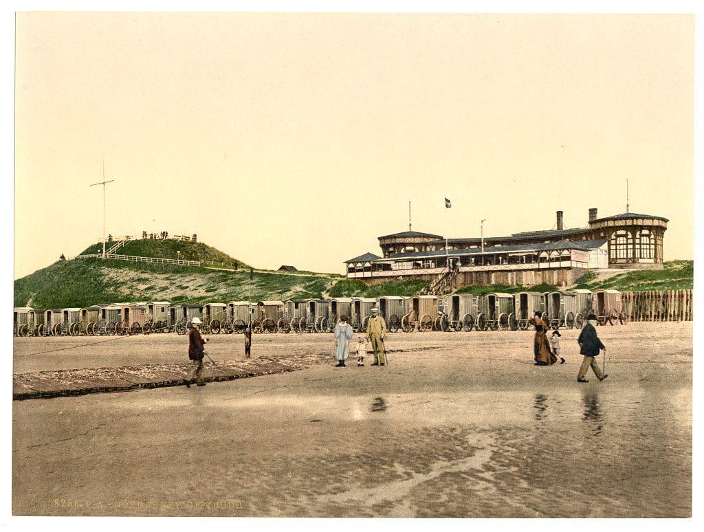 The image depicts a historical scene likely from the late 19th century. In the foreground, there are several individuals walking along what appears to be a boardwalk or sandy beach area near water with visible puddles reflecting light on their surfaces. They are dressed in clothing indicative of that era's fashion, suggesting leisure activities.

In the middle ground, rows of horse-drawn carts and wagons stretch across an open field leading towards a large building perched atop a small hill to the left. The structure has multiple levels with various dormer windows and appears to be designed for public use or recreation. A flag is flying above it on what seems like a pole.

The background features more of these horse-drawn vehicles, suggesting this could be an event, market, or transportation hub at some distance from urban centers during that time period.

Overall, the scene captures daily life with elements of tourism and commerce in Germany around 1890-1900.