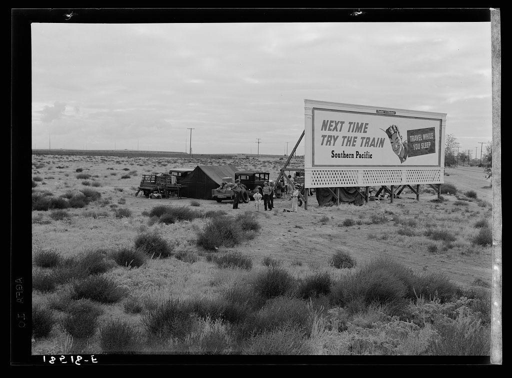The image depicts a black and white photograph taken on U.S. Route 99 in California, showing three families who are living under the open sky with their belongings spread out around them for protection from wind or sun. They appear to be camped behind a billboard that serves as a partial windbreak.

In this scene, there is an emphasis on outdoor life and hardship during what seems like difficult times. The photograph captures a moment in history where people were possibly displaced due to droughts, the Great Depression, or war-related evacuations.

The black and white photo adds to its historical context by illustrating how such conditions might have been visually represented at that time period.