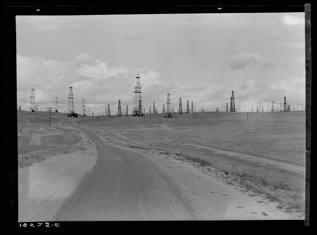 The photograph depicts a vast oil field landscape with numerous tall drilling rigs scattered across the horizon. The sky is partly cloudy, and there's an expansive dirt road leading into the distance on the left side of the image. This black-and-white photo exudes a vintage quality, indicative of early to mid-20th-century photography techniques. It captures the desolate yet industrious atmosphere characteristic of oil extraction sites during that era in California, particularly Kern County.