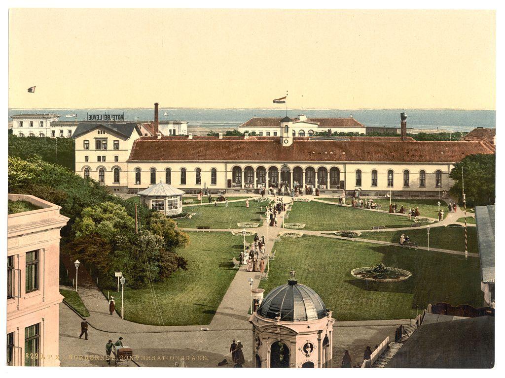 The image depicts a grand building with classical architecture, featuring rows of arched windows and prominent columns along its facade. The structure is surrounded by well-maintained gardens with neatly trimmed lawns, flowerbeds, and pathways where people are seen strolling or sitting on benches. A central fountain adds to the aesthetic appeal of the scene. In the foreground stands a dome-topped building with classical details, possibly serving as an entrance or pavilion for visitors. The overall atmosphere conveys elegance and leisurely enjoyment in a European park-like setting during what appears to be late 19th-century Europe.